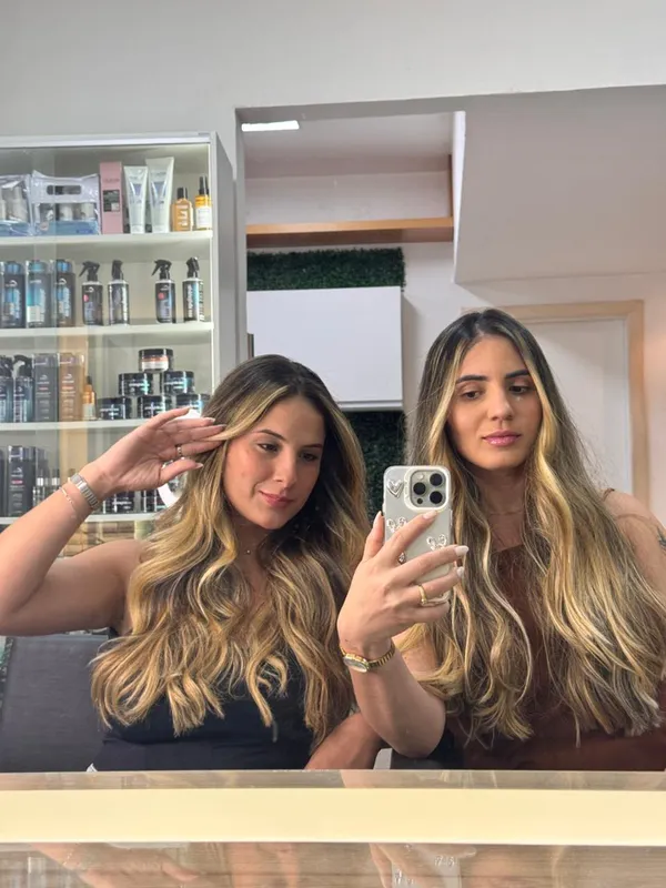 Duas mulheres com cabelo longo ondulado e mechas loiras douradas, fazendo selfie no espelho do salão