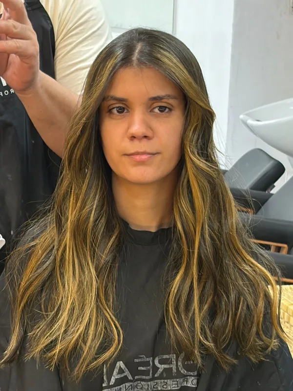 Mulher de frente com cabelo longo castanho escuro e mechas douradas, sentada na cadeira do salão