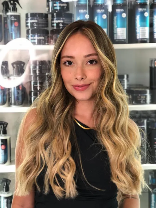 Mulher sorrindo com cabelo longo ondulado e mechas loiras douradas diante de prateleiras de produtos