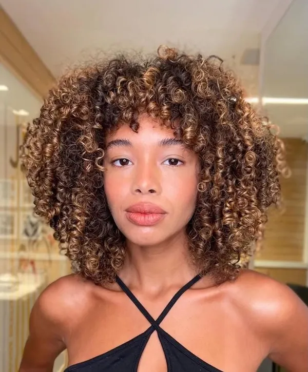 Mulher de frente com cabelo cacheado volumoso, franja e mechas em tom caramelo