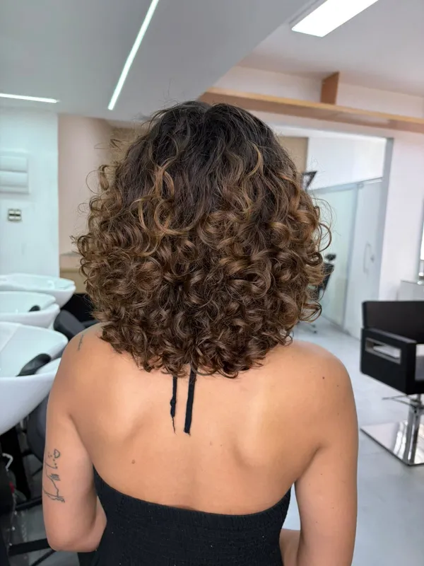 Mulher de costas com cabelo cacheado na altura dos ombros, raiz escura e pontas em tom caramelo