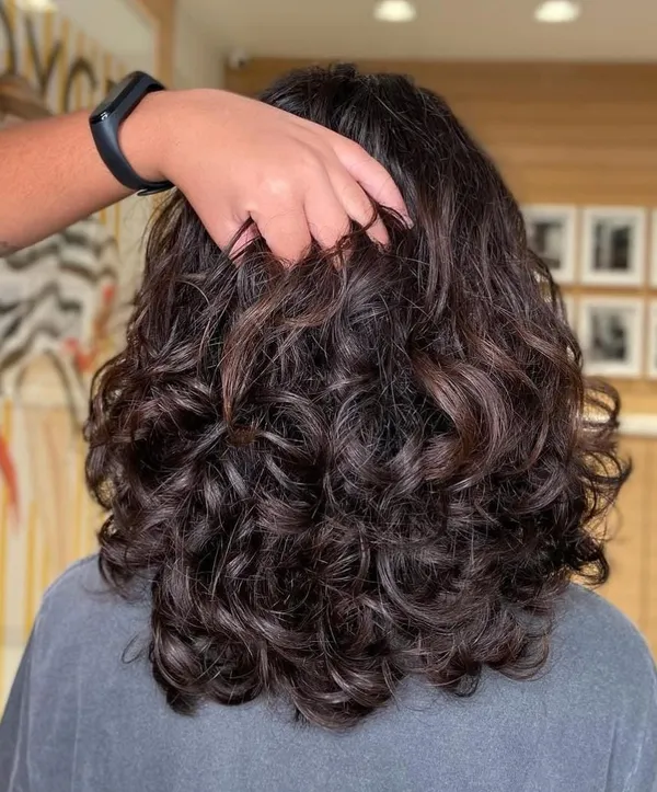 Cabelo castanho escuro em cachos largos e volumosos, visto de costas, com a mão do cabeleireiro sobre a cabeça