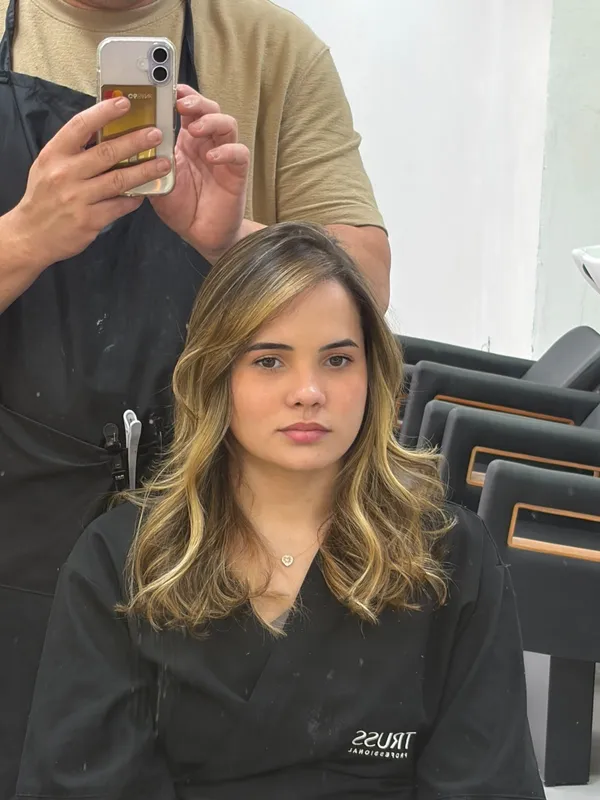 Cliente sentada com cabelo com mechas douradas, enquanto o cabeleireiro fotografa o resultado no espelho