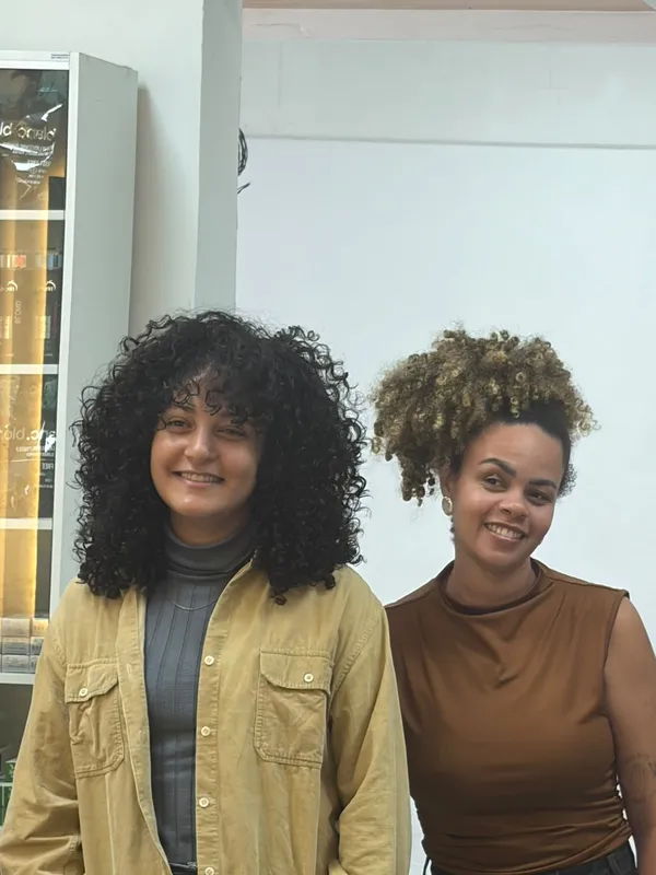Duas mulheres sorrindo, uma com cabelo cacheado escuro e volumoso e outra com cabelo crespo com mechas douradas