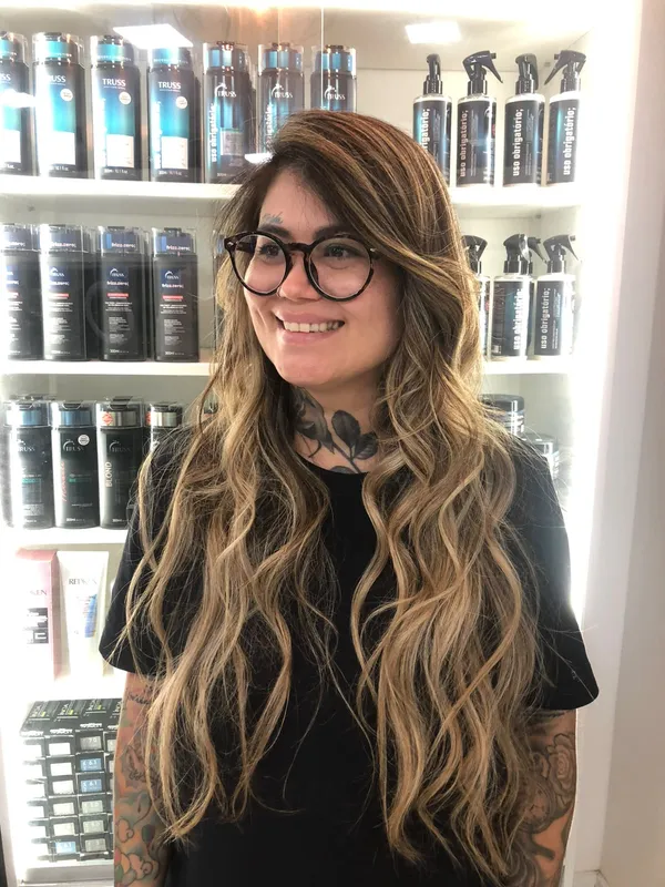 Mulher sorridente com óculos e cabelo longo ondulado com mechas douradas, diante de prateleira de produtos
