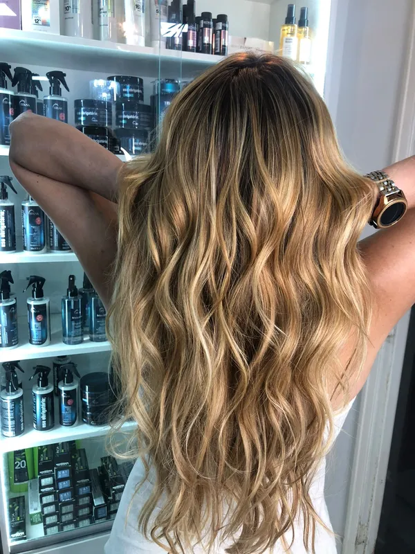 Mulher de costas com cabelo longo ondulado e mechas douradas, com as mãos atrás da cabeça