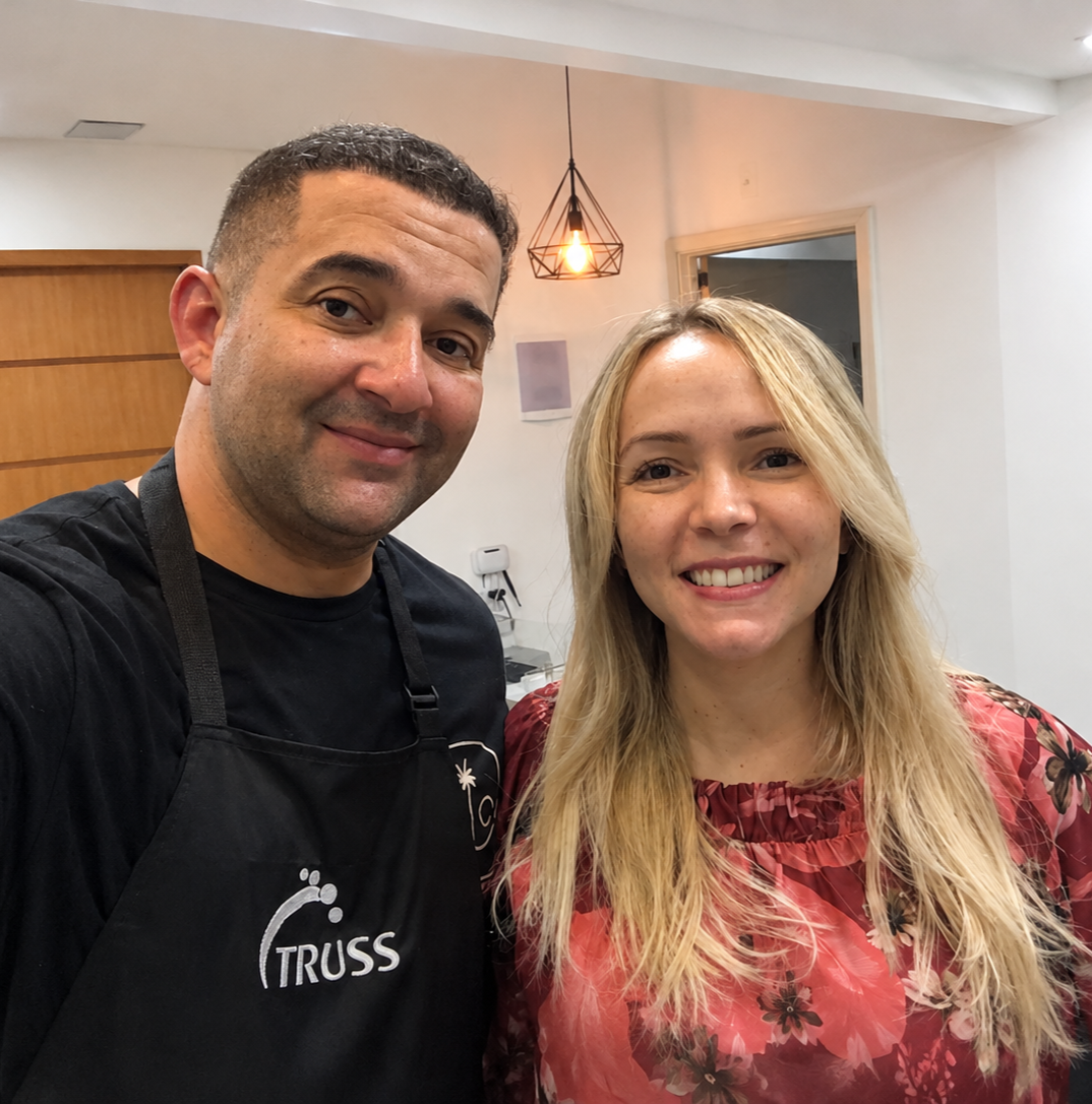 Flávio Barcelos, cabeleireiro, de avental preto, ao lado de uma cliente sorrindo.
